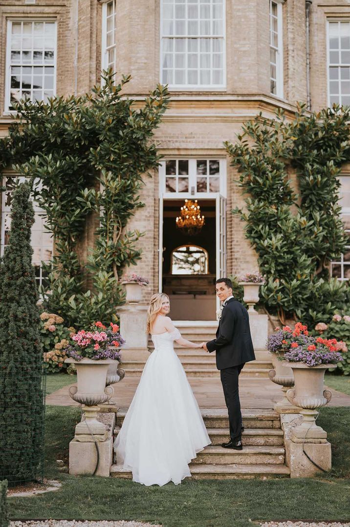 Bride in off shoulder wedding dress with lace detail and groom in black tux holding hands outside Hedsor House