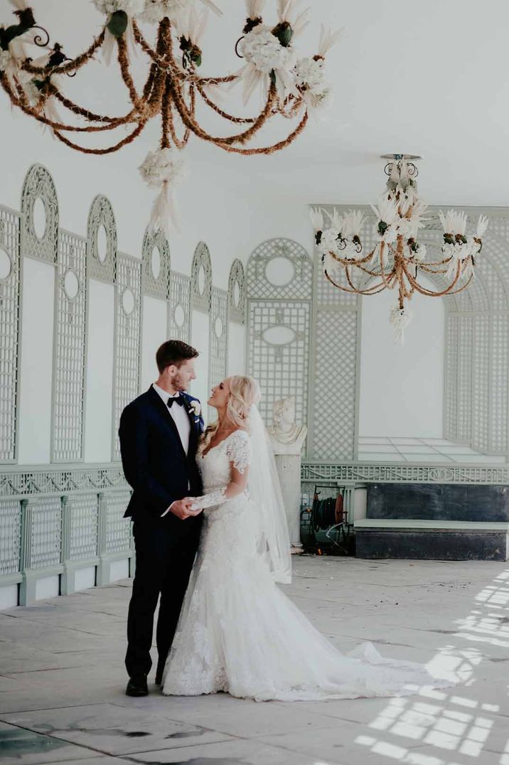 Bride in lace wedding dress and groom in classic tux standing in Port Eliot Estate with large gold chandeliers 