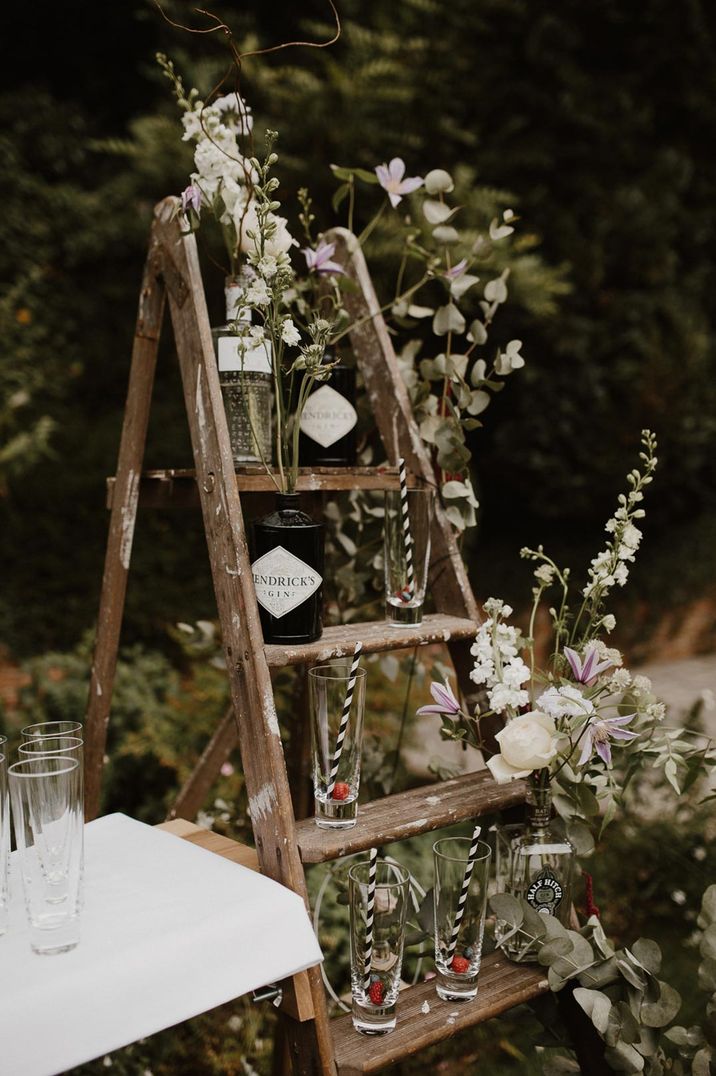 Wooden ladder with DIY wedding gin bar - Hendrick's bottle, tall glasses with stripes straws and foliage and floral decorations wrapped around 