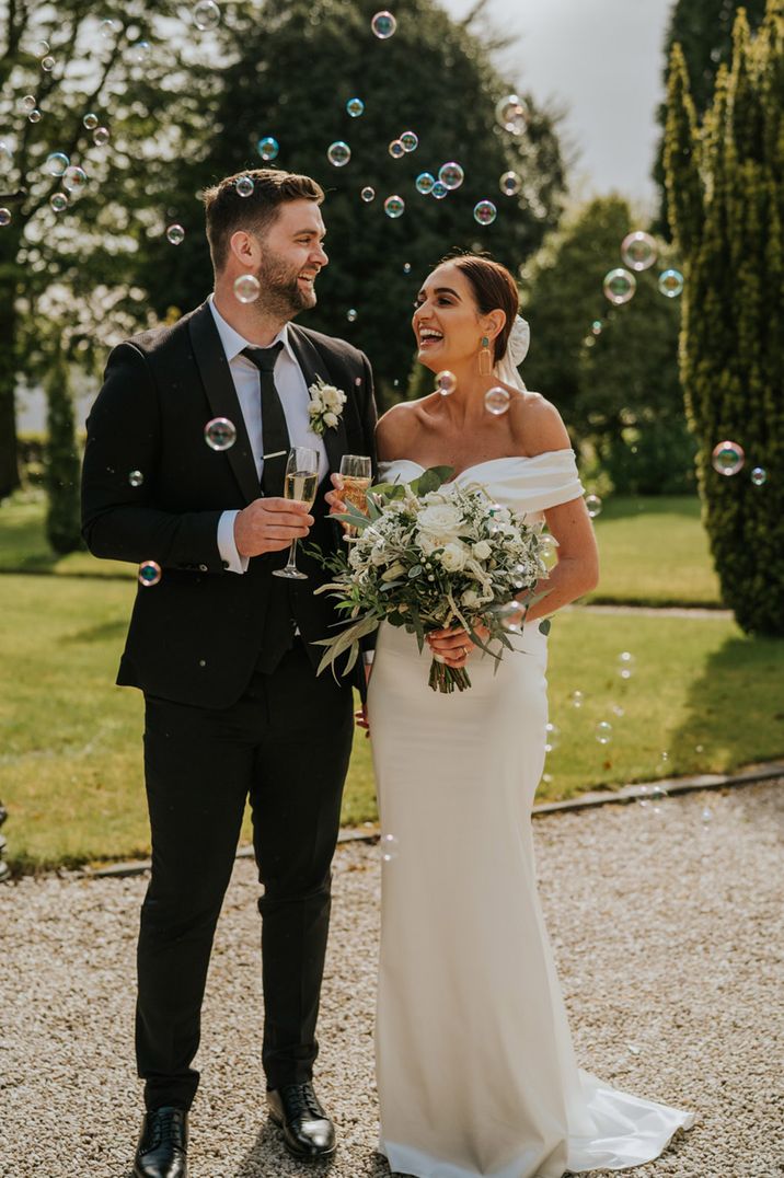 Bride in off the shoulder wedding dress with the groom in a black tuxedo drinking a glass of champagne surrounded by bubbles 