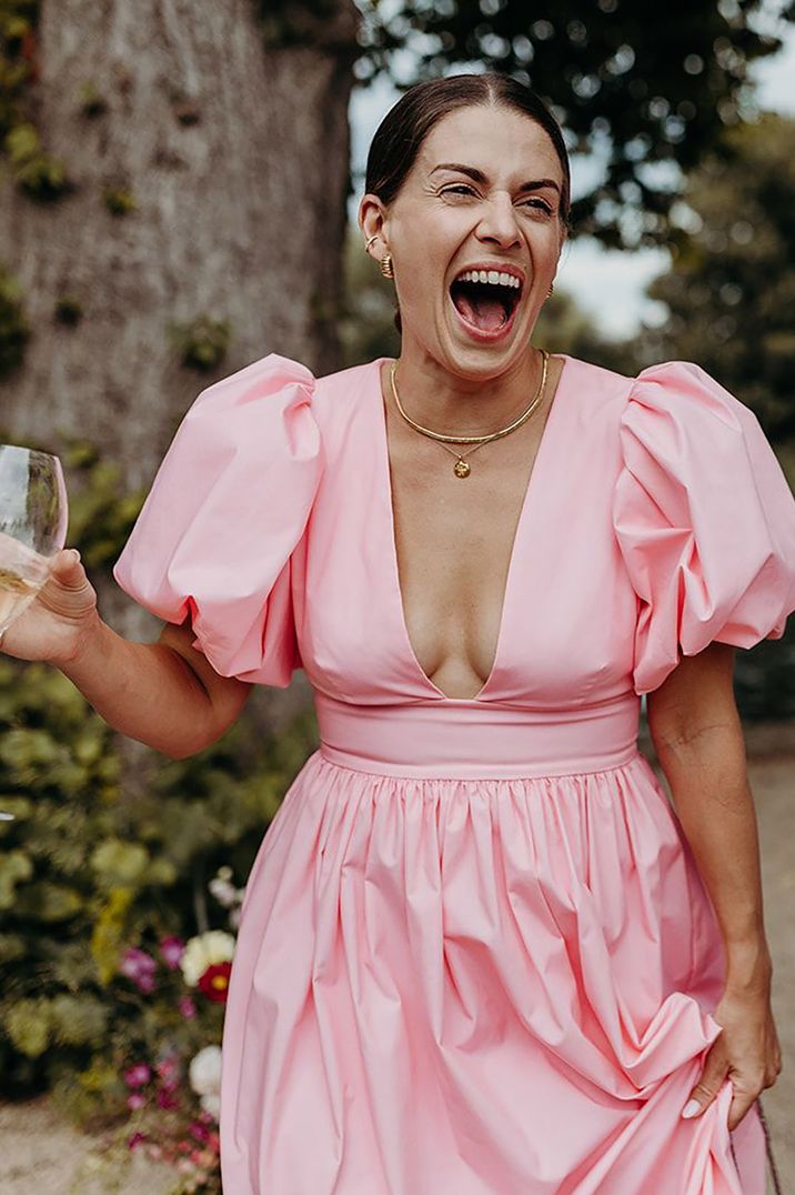 Wedding guest wearing a pink summer wedding guest dress smiling widely holding a glass of bubbles by Richard Skins Photography