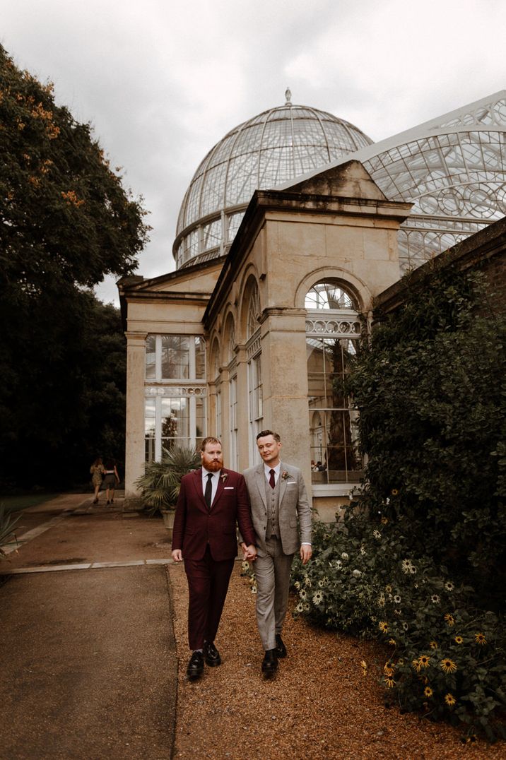 Groom in a burgundy suit holding hands with his husband in a beige check suit outside Syon Park wedding venue 