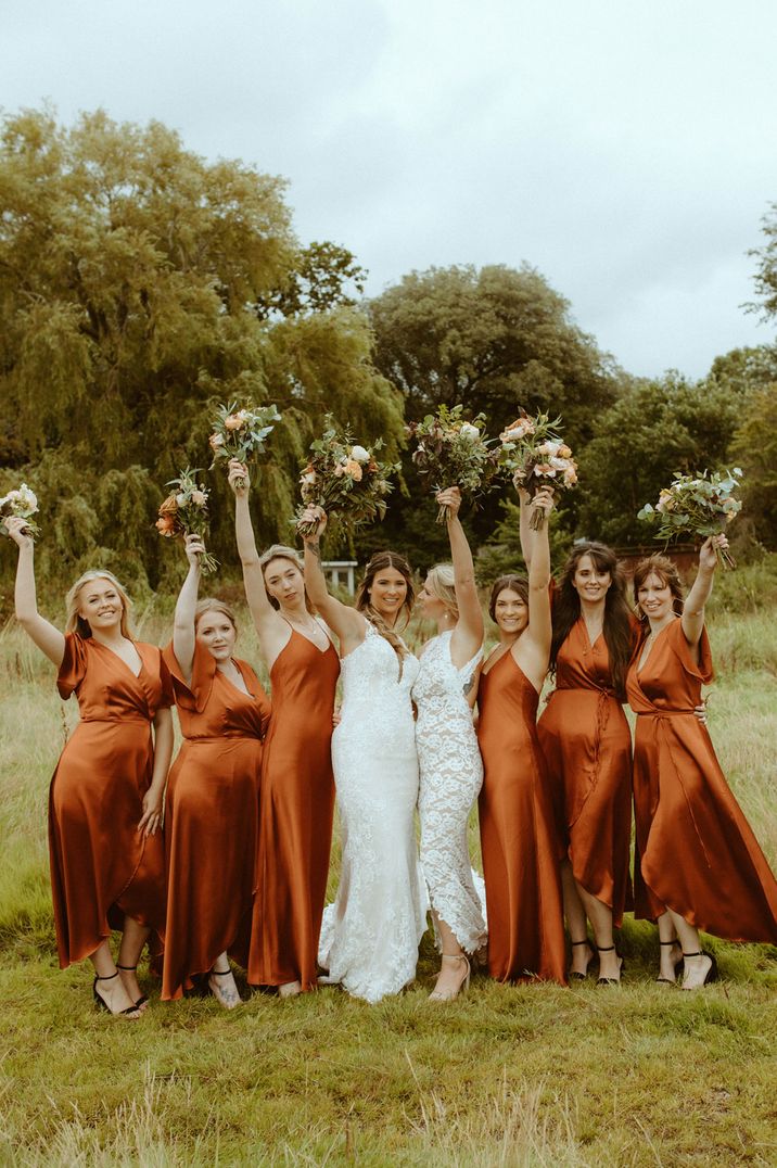 Two brides in bridesmaid dresses stand holding up bouquets with bridesmaids in burnt orange satin bridesmaid dresses