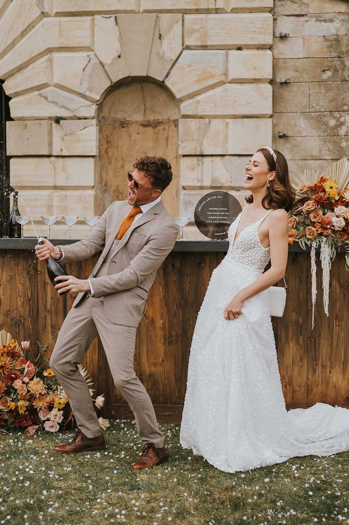 Bride in an appliqué wedding dressing groom in a beige check suit popping champagne 