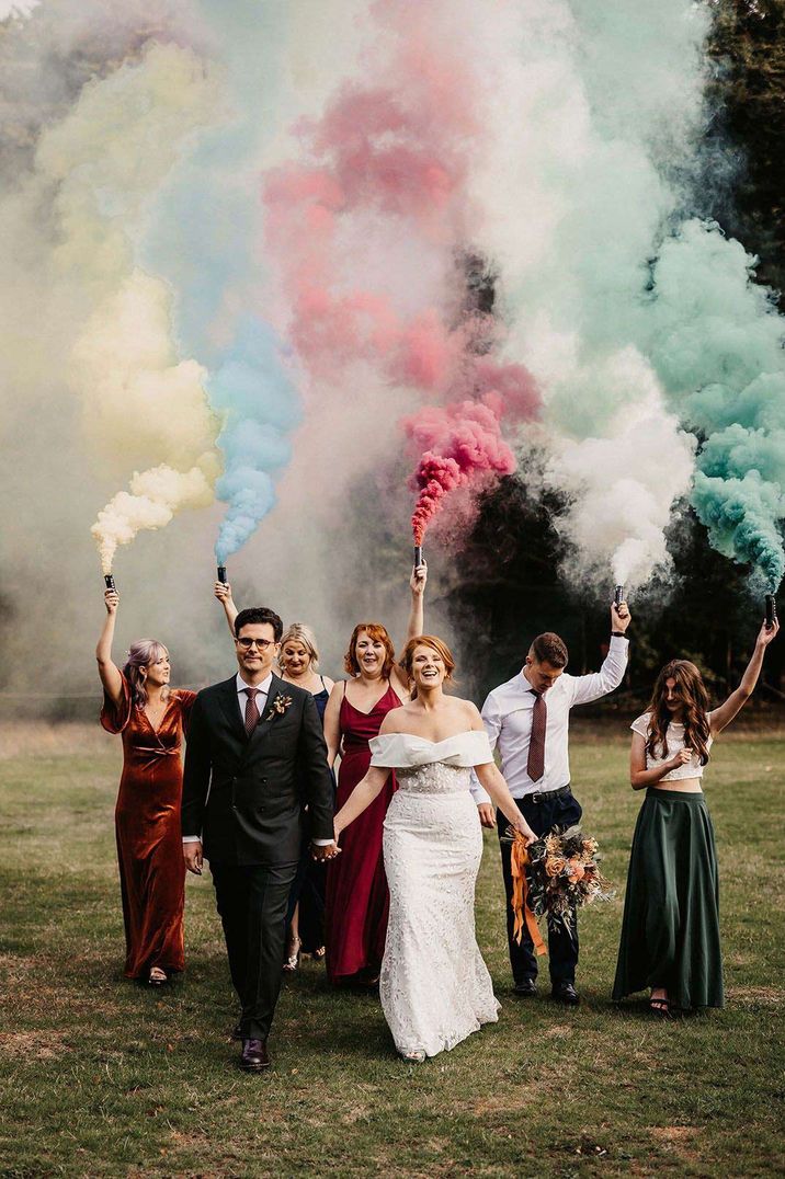 Bride in off the shoulder lace wedding dress and groom in classic tuxedo doing colourful smoke bomb photoshoot with wedding party 