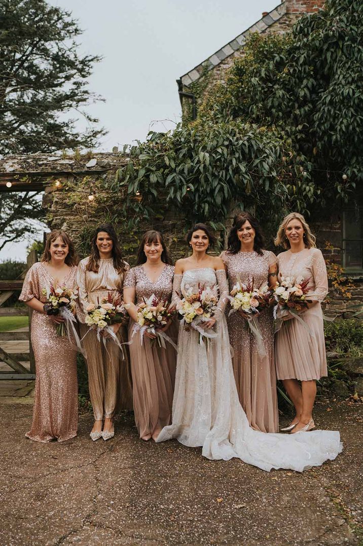 Bride in off shoulder embellished wedding dress standing with bridal party wearing mismatched gold embellished bridesmaid dresses 