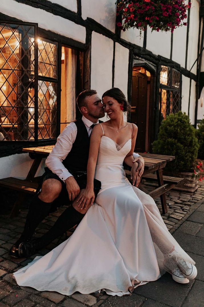 bride in a Stephanie Allin wedding dress with front slit sitting on her grooms lap outside their pub reception 