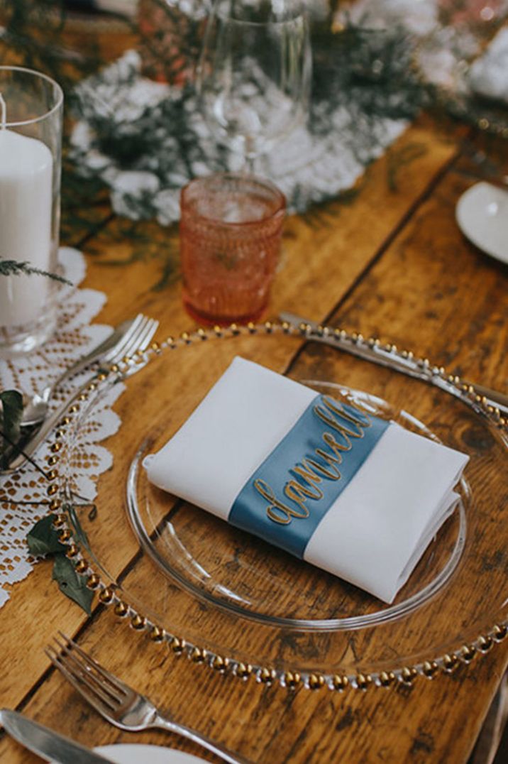 Clear charger plate with gold with the white napkin placed in the middle topped with a blue ribbon and gold place name 