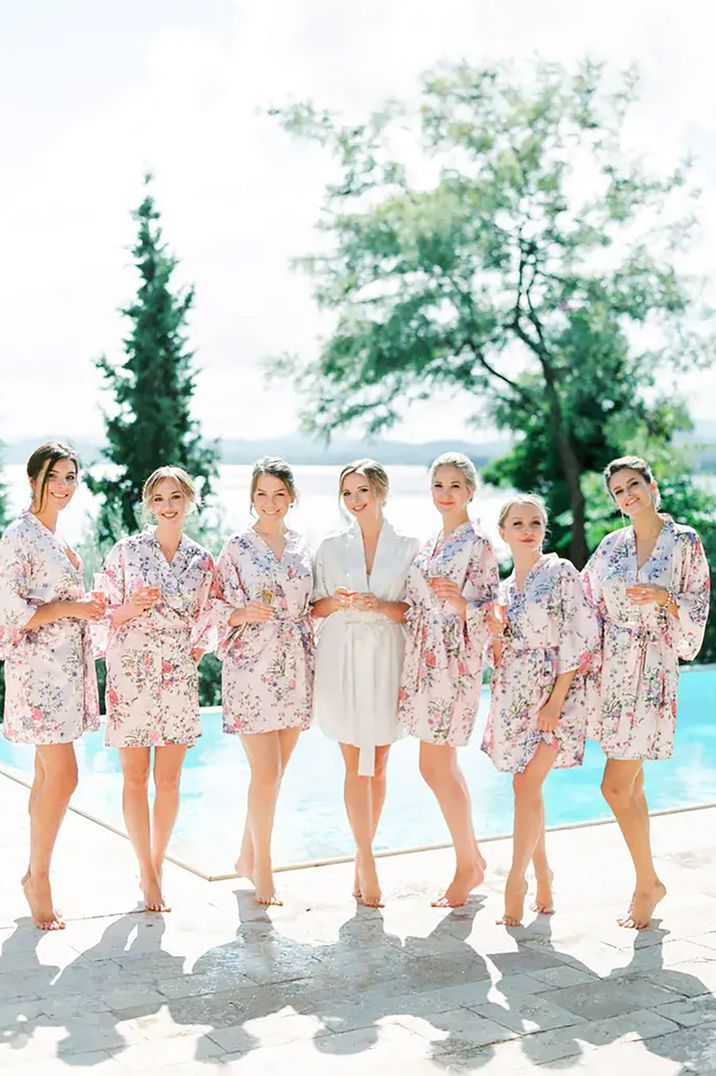 Bride and her bridesmaids standing in a line posing for an image by an outdoor pool wearing robes by Mikhail Balygin Photography