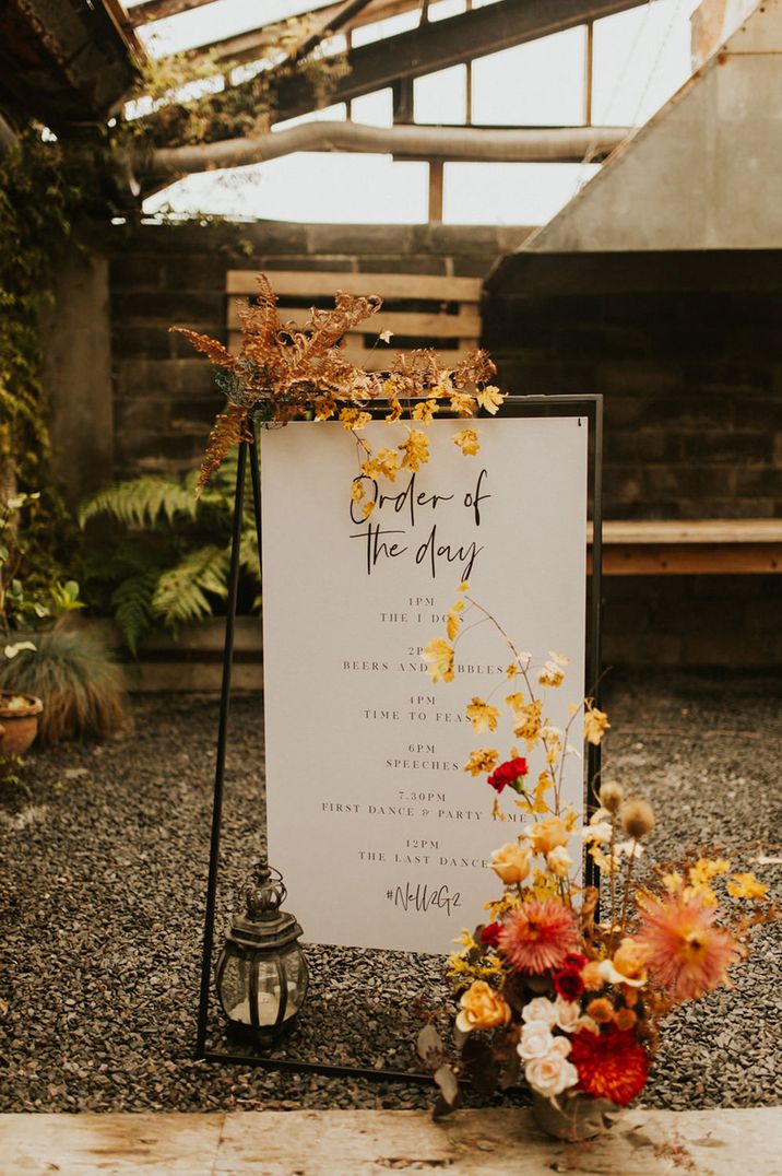 Order of The day wedding sign on a black metal frame decorated with orange autumn flowers and leaves