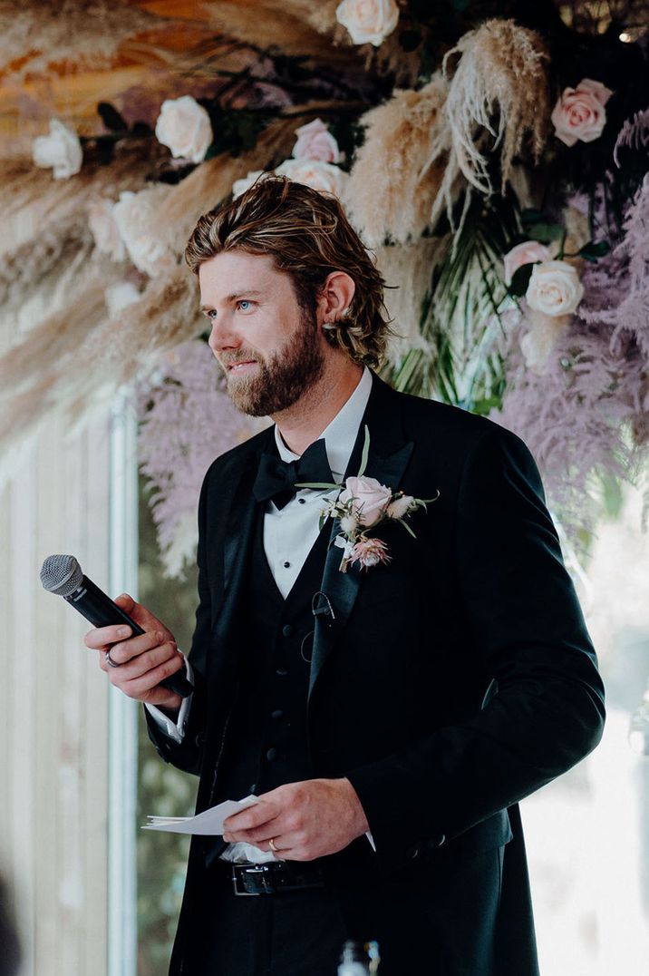 Groom in black tux and neutral boutonnière giving speech at wedding