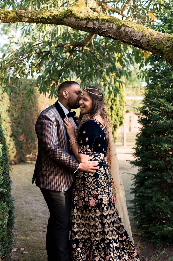 Woman in intricate outfit with gold jewellery and headpiece getting kissed by the groom in a patterned suit  