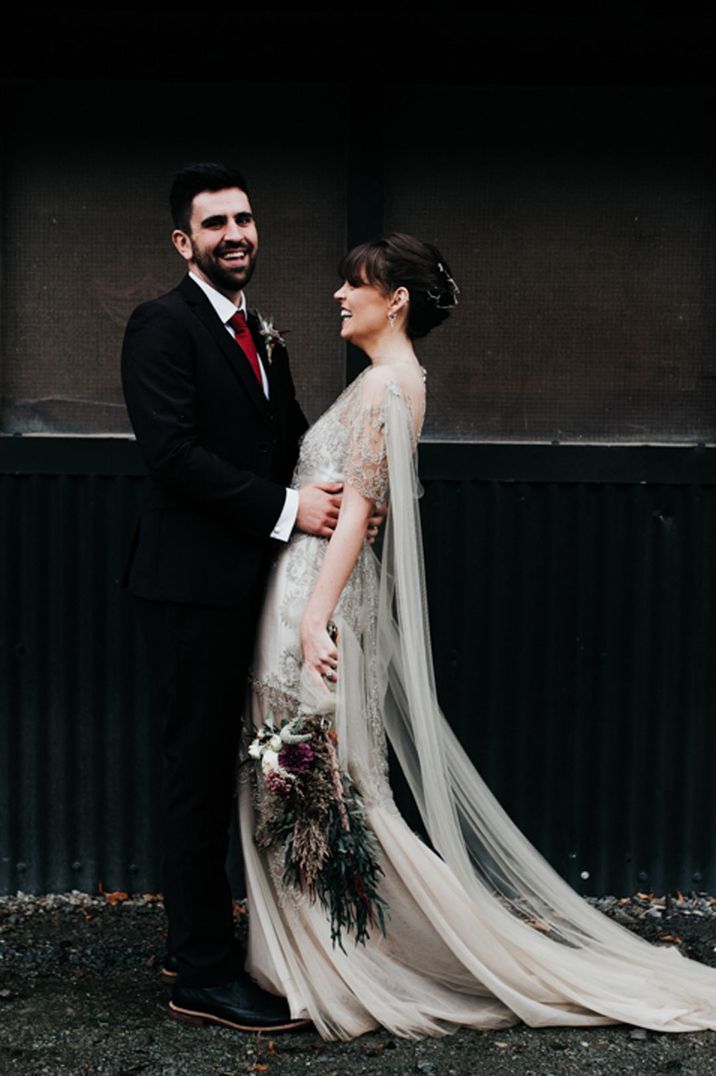 Bride in homemade sage tinted quarter length sleeve wedding dress with homemade bouquet and peacock feathers and groom in dark suit with red tie