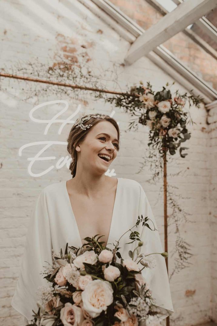 Bride in v neck long sleeve wedding dress holding neutral luxe rose, peony and foliage bouquet standing in front of rose gold frame and white light neon wedding sign 