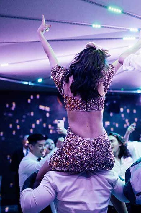 Bride in sparkly two piece bridal set and groom in white shirt and black trousers dancing on guests' shoulders 