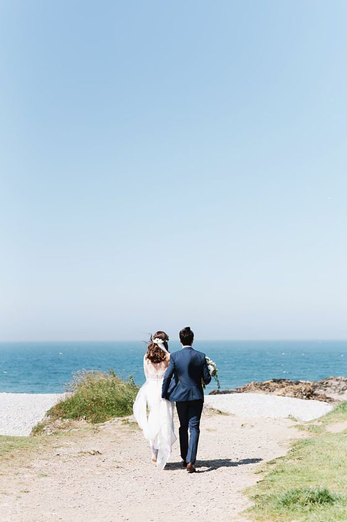 Bride and groom walking towards the sea for beach wedding with bride wearing a lace low back beach wedding dress