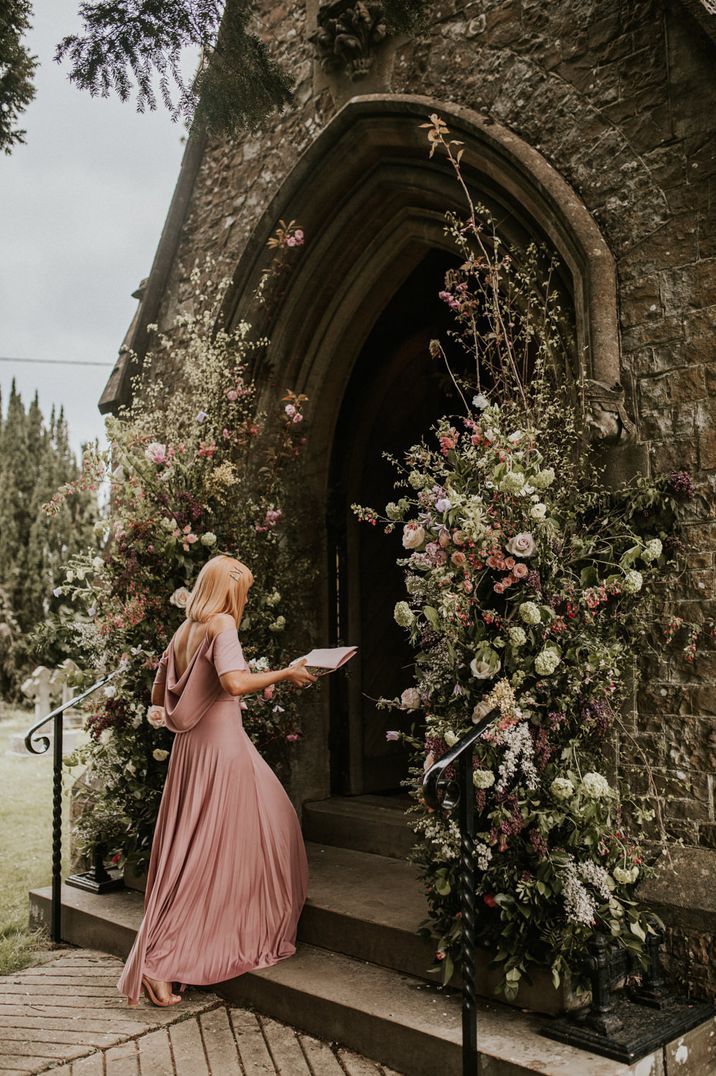 Bridesmaid in a pink cowl back dress walking into the church decorated with wild flower arrangements