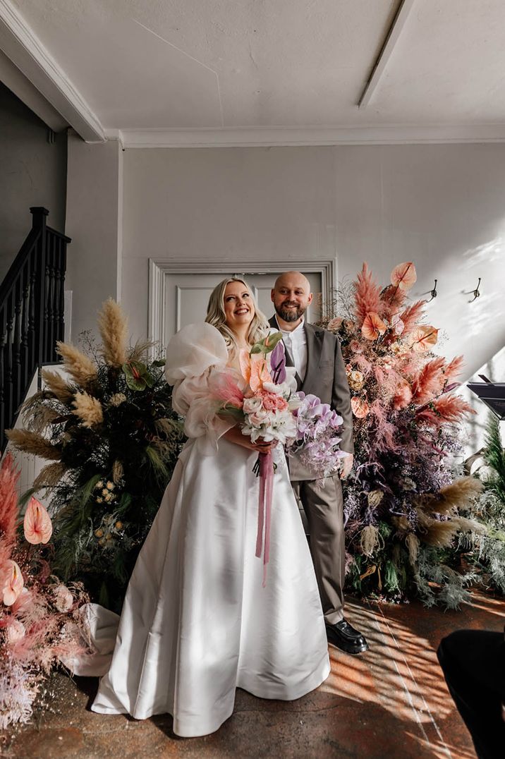 Bride and groom smile in front of flower columns with pastel coloured pampas grass 