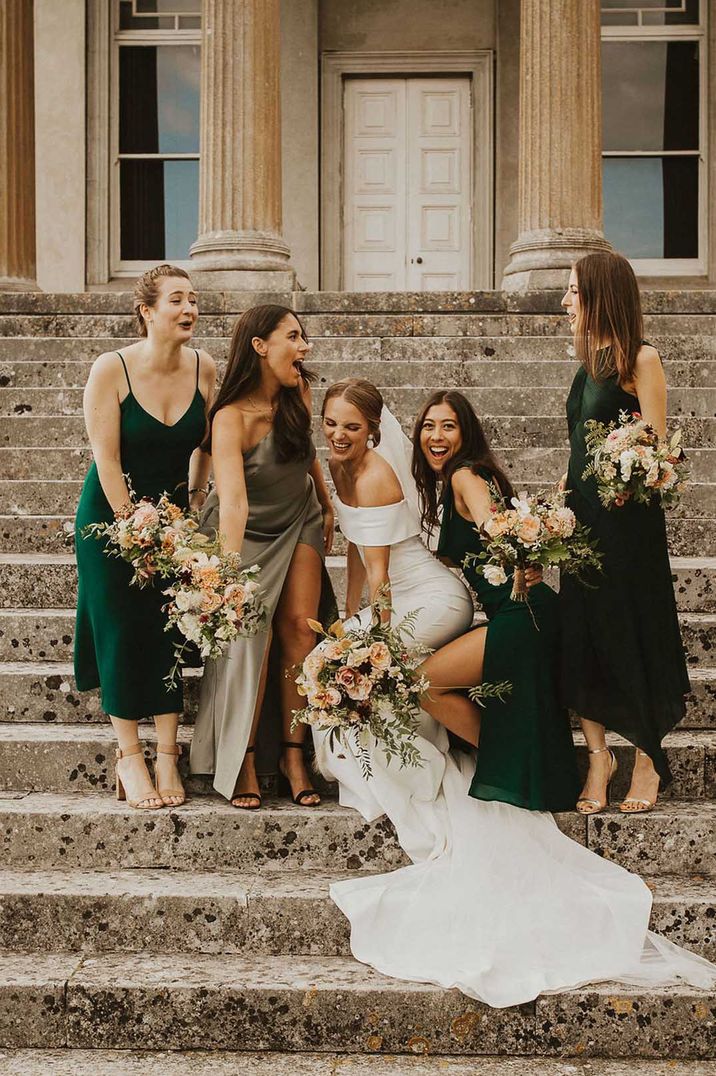 Bride in off the shoulder bodycon wedding dress standing on the steps of The Grange Hampshire with bridesmaids in mismatched green bridesmaid dresses 