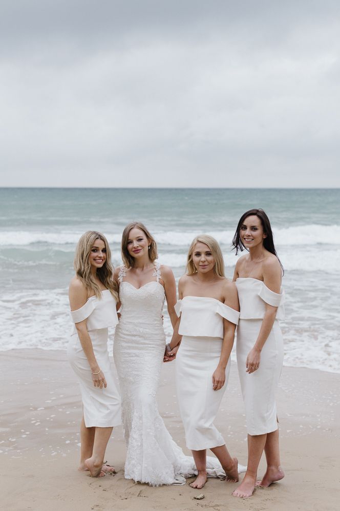 Bridal party standing and posing on the beach wearing midi white bridesmaid dresses