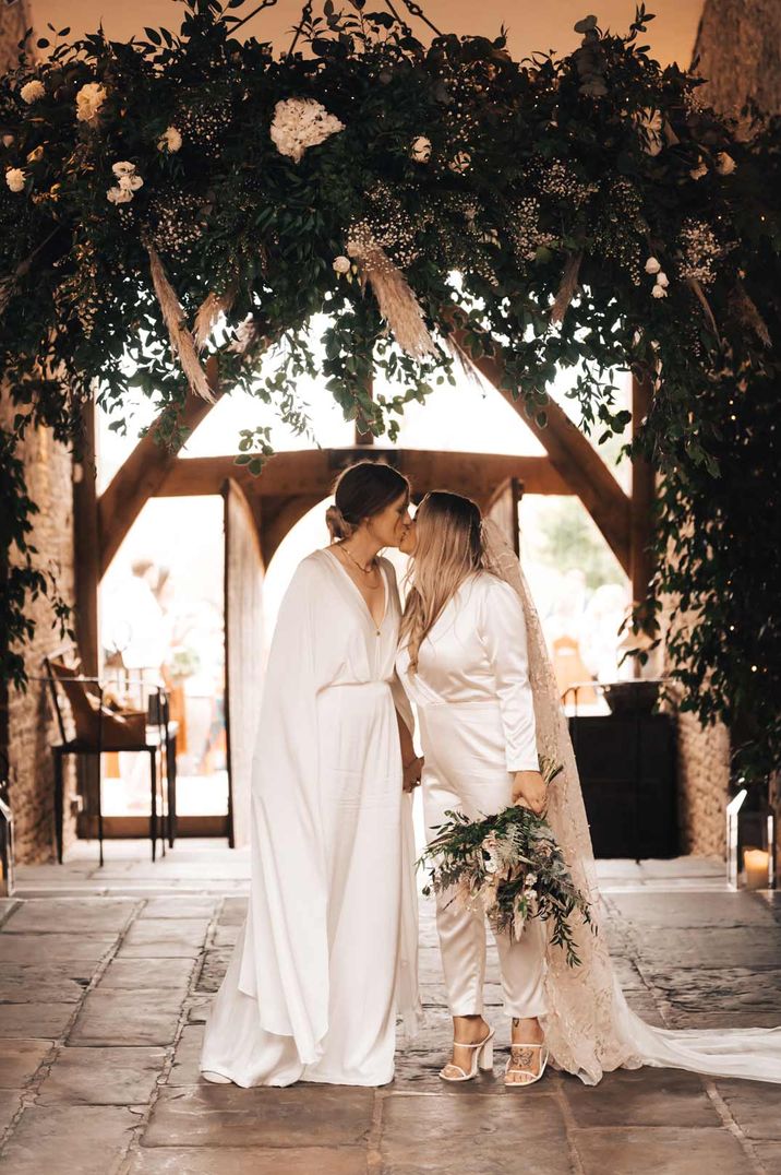 Two brides in complimenting white bridal jumpsuits embracing at rustic wedding venue