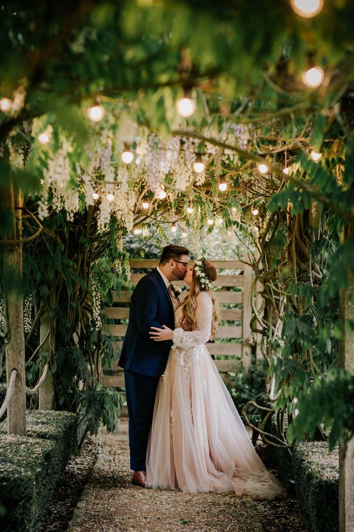 Bride in long sleeve lace wedding dress and groom in tux embracing amidst greenery and foliage of barn wedding venue