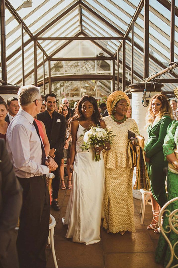 Bride in sleeveless satin wedding dress walking down the aisle with mother of the bride in yellow polka dot outfit 