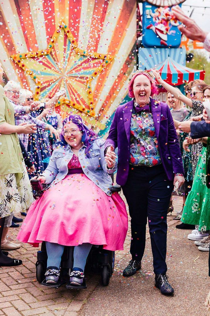 Bride in bright pink wedding dress and personalised denim jacket doing colourful confetti exit with bride in deep purple velvet blazer 