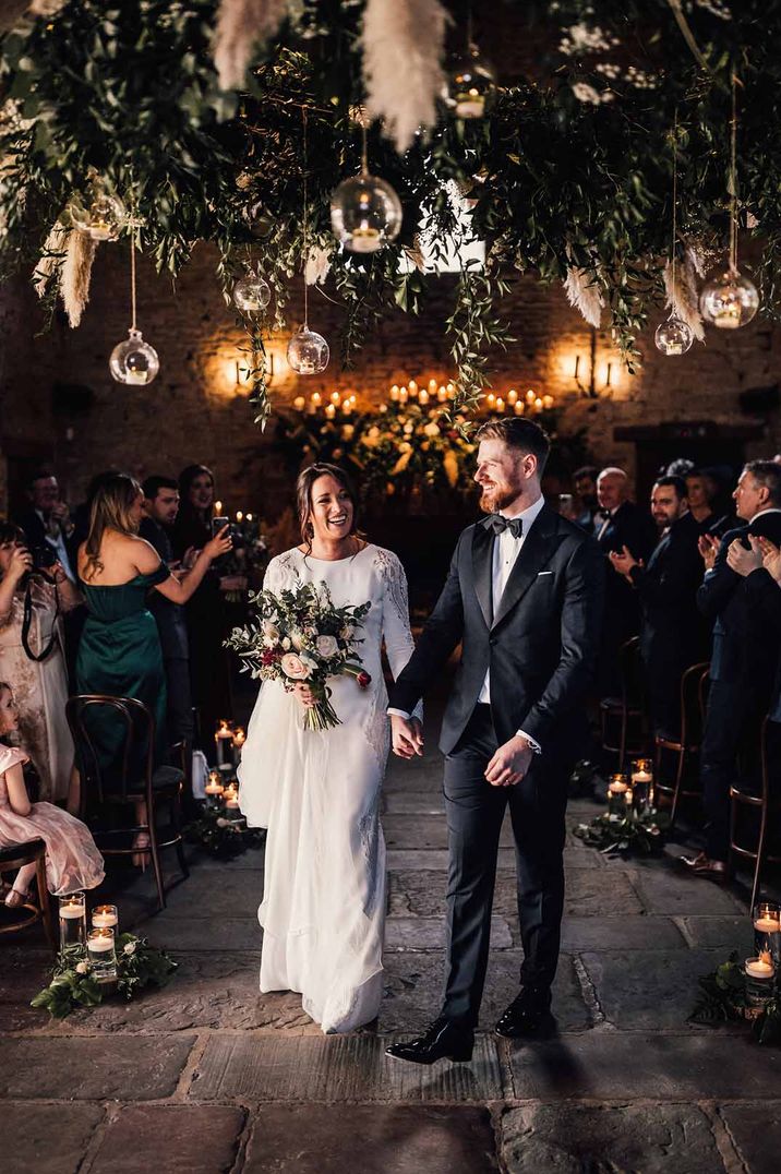 Bride in long sleeve wedding dress walking with groom in classic black tuxedo in Cripps Barn with disco ball and pampas grass wedding decor suspended 