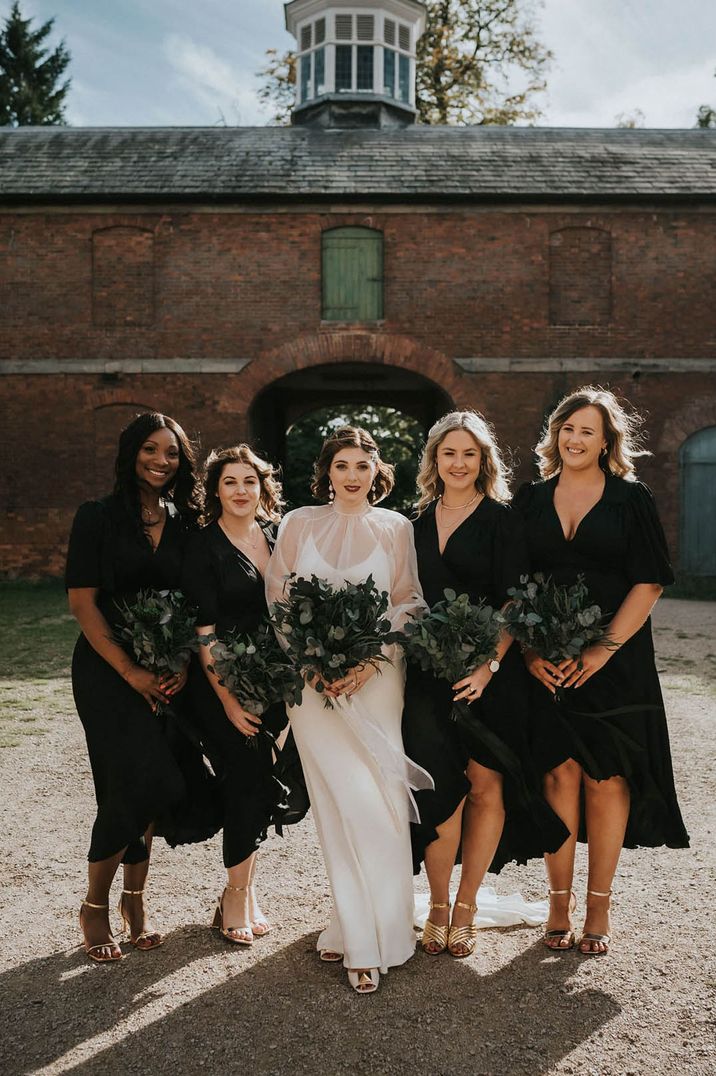 Bridal party shot of bridesmaids wearing black dresses and gold metallic shoes sanding with the bride in a sheer long sleeve wedding gown holding all foliage preserved bouquets 