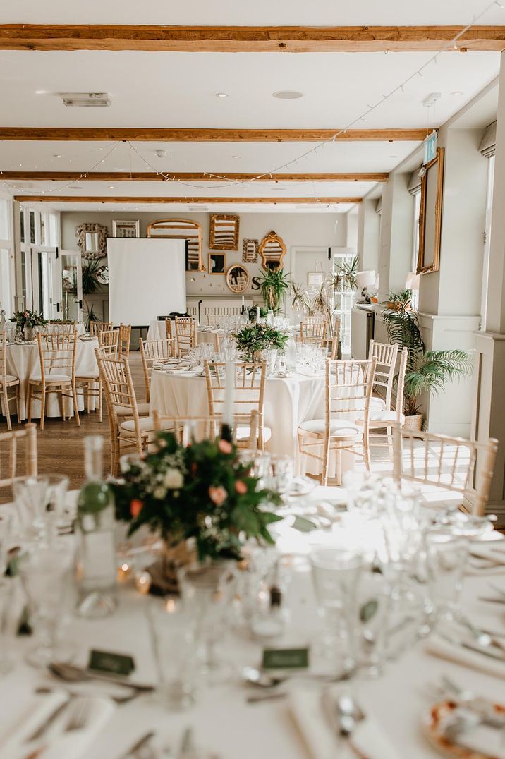 Neutral wedding tablescape with white tablecloths, foliage and rose wedding centrepieces, wooden beams and fairy lights