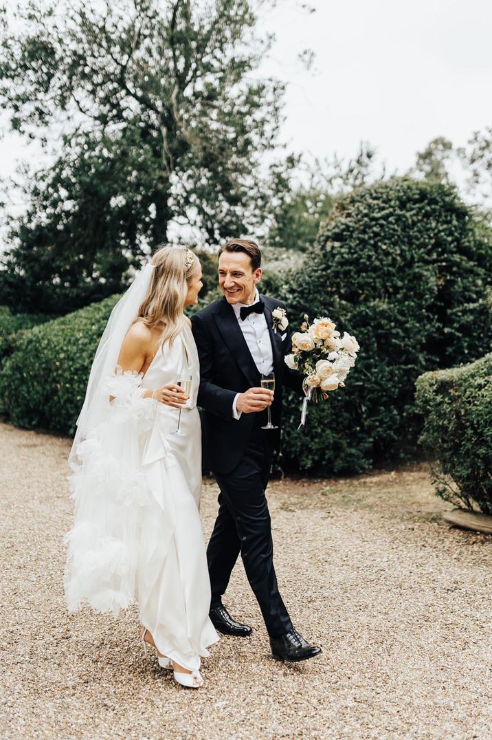 Groom in a black tuxedo walking with the bride in a halter neck wedding dress with feathers for traditional wedding 