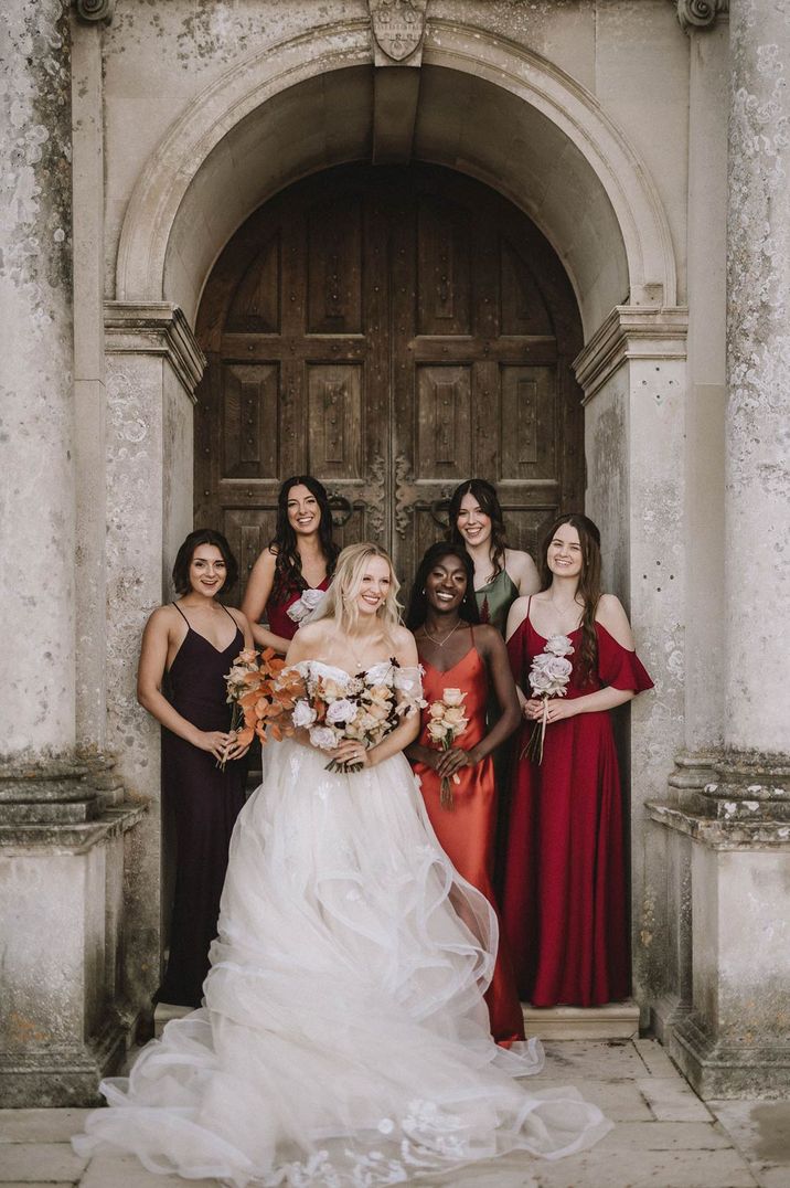 Bride in off shoulder princess wedding dress standing with bridesmaids in satin mismatched bridesmaid dresses 