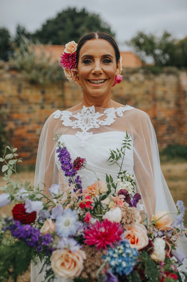 Bride in wedding dress with cape holding colourful wedding bouquet with flower hair accessories for vow renewal wedding 