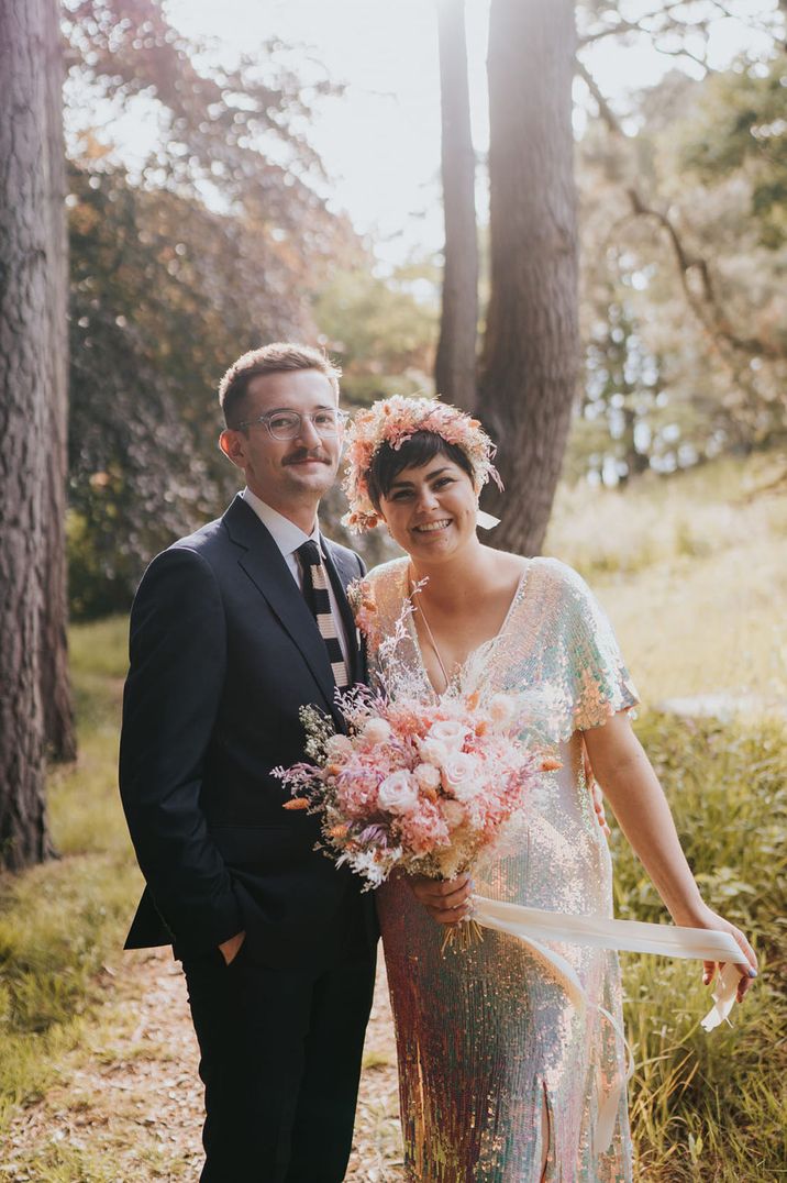 Bride in colourful sequin wedding dress with flower crown and lush preserved pink flower bouquet tied with pink ribbon standing with the groom in a navy suit and striped tie