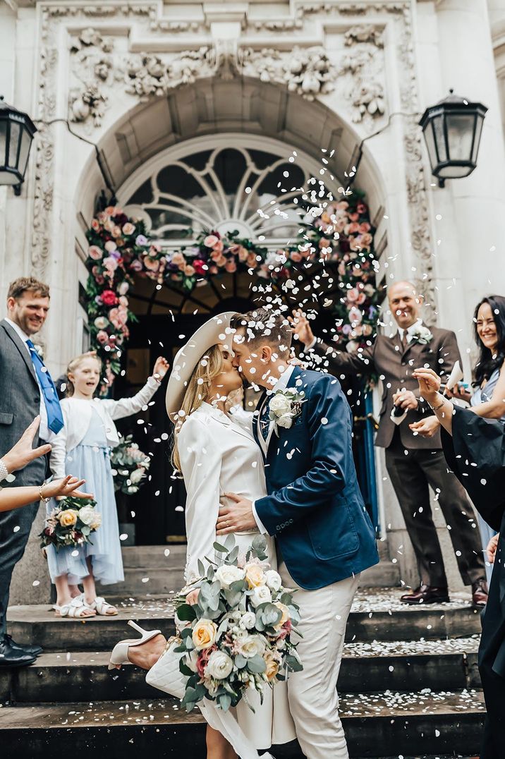 Bride in long sleeve wedding dress and bridal hat kissing groom in blue blazer doing confetti throw - how to save for your wedding