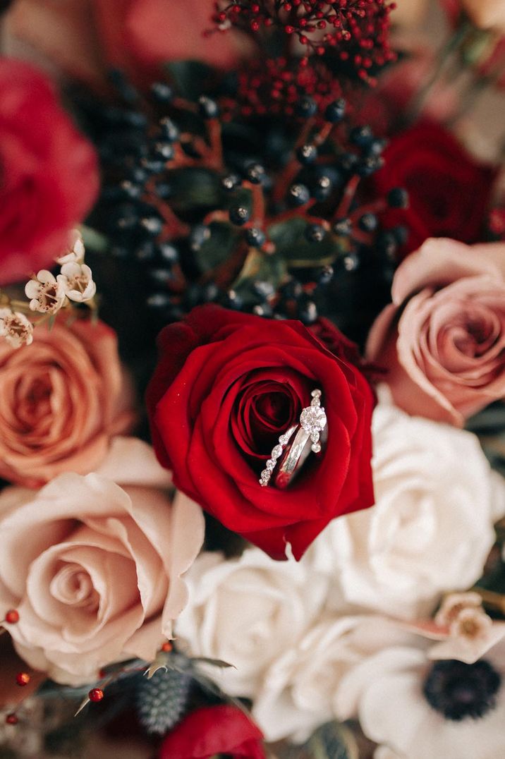 Bouquet of red roses and other red flowers with diamond round brilliant engagement ring and wedding bands for the bride and groom 