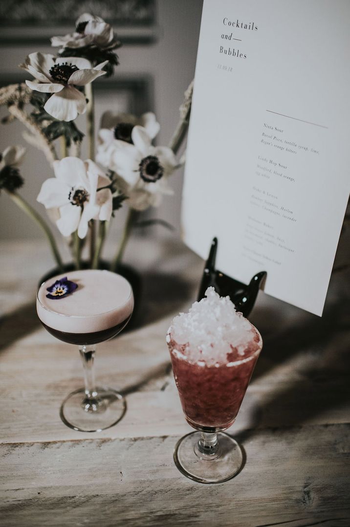 Glass with crushed ice and red cocktail next to another glass of foamy cocktail with edible flowers 