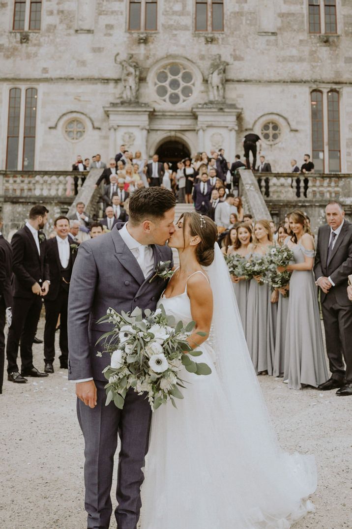 Groom in a grey suit with white tie kissing his bride in a cami top wedding dress with tulle skirt outside Lulworth castle 