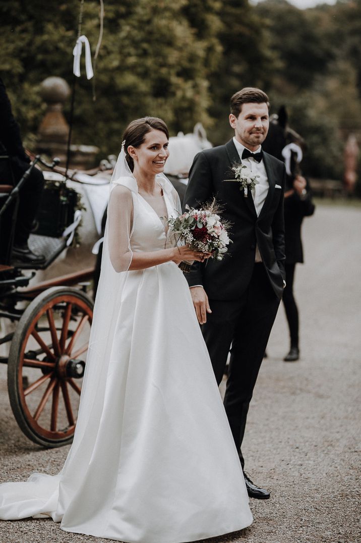 Bride in long sleeve sheer wedding dress and groom in black tie in front of wedding carriage