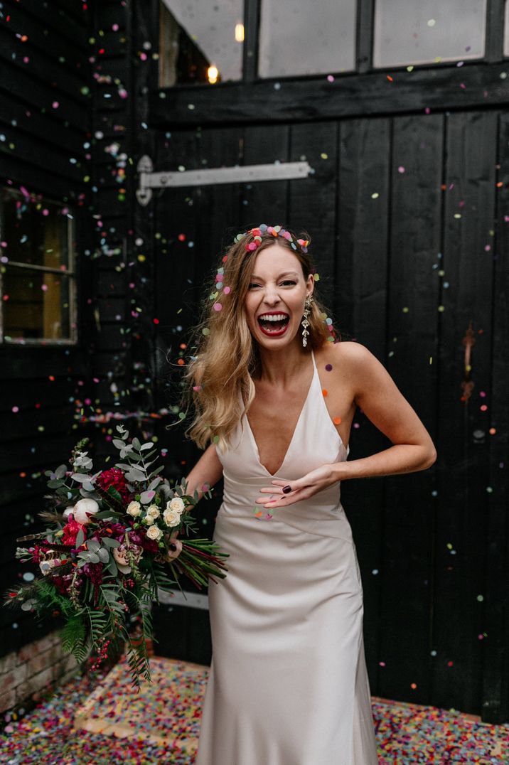 Bride in a satin wedding dress with a red lip smiles as colourful confetti explodes 