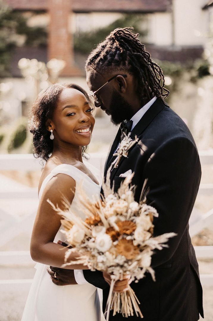 Bride in off the shoulder wedding dress smiling at the camera with the groom in black tie holding dried flower pampas grass bouquet 