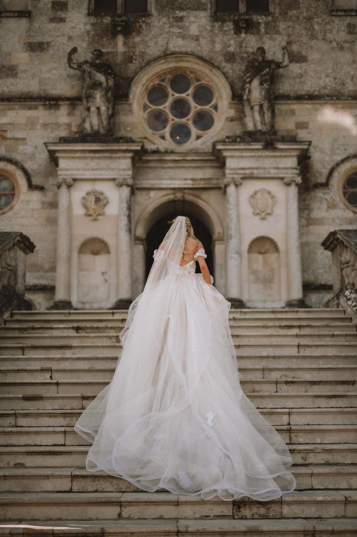 Bride in large off the shoulder princess wedding dress with tulle finish walking up the steps of Lulworth Castle 