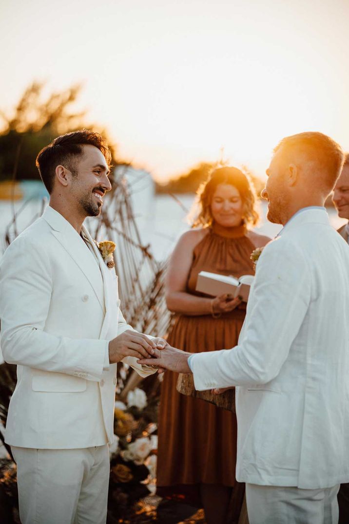 Golden hour wedding portrait with two grooms in white wedding suits exchanging vows on the beach 