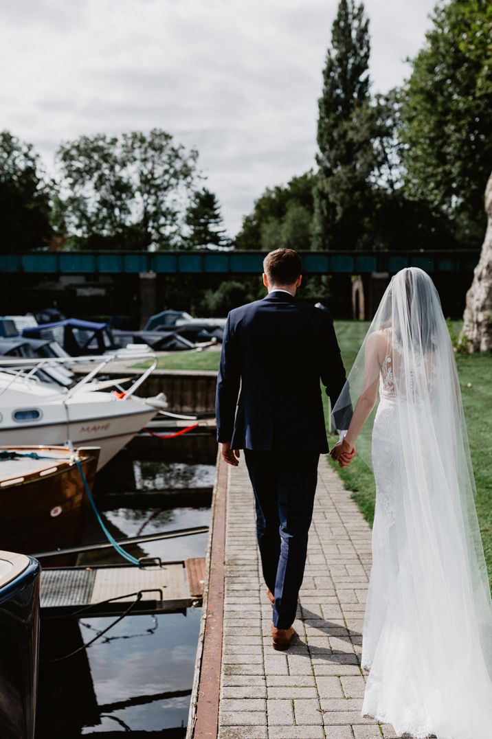 Bride in a lace wedding dress with cathedral length veil holding hands with her groom by the canal 