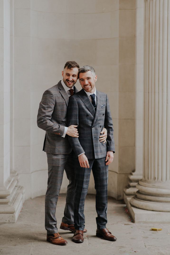 Grooms stand with one another outside Old Marylebone Town Hall in front of white pillars on their wedding day 