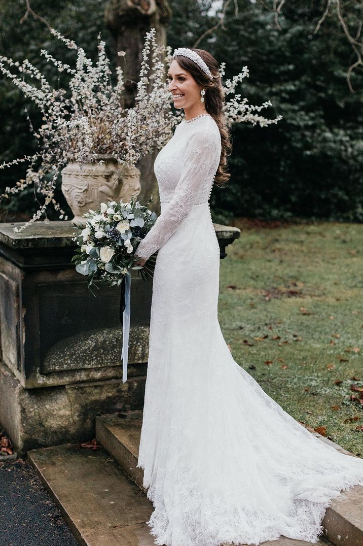 Bride in a lace long sleeve wedding dress with a white flower bouquet, pearl headband and statement earrings