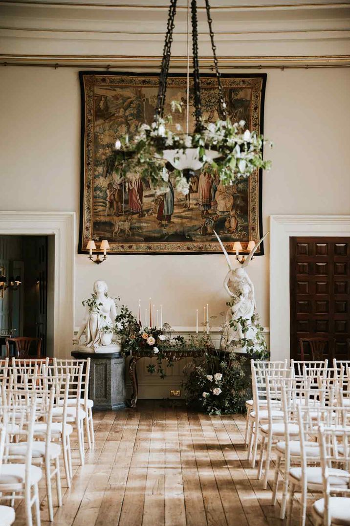 Light, natural ceremony room at Elmore Court wedding venue with festoon chandelier lighting, white rose and foliage decor, two angel statues and tapered candles; how to book wedding venue