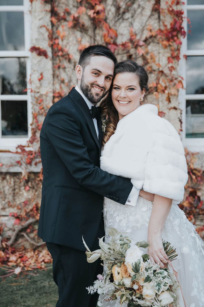 Bride in lace wedding dress and faux fur shawl posing with the groom in a black tuxedo for winter wedding 