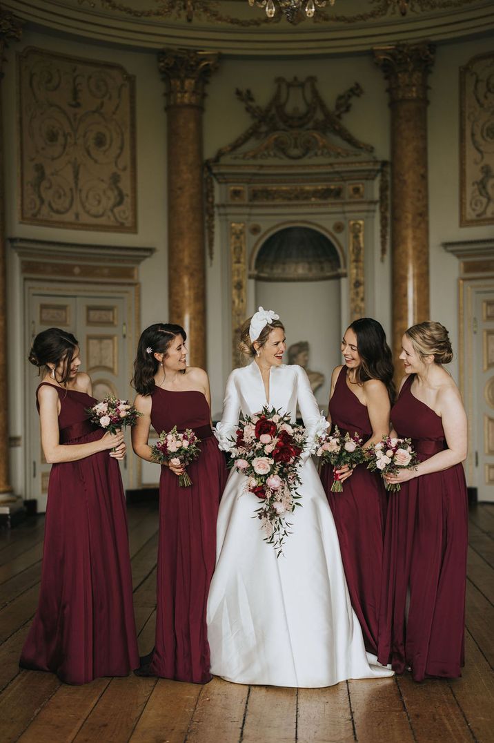 Bride wearing classic long sleeve satin wedding dress standing with bridal party wearing one sleeve burgundy bridesmaid dresses 